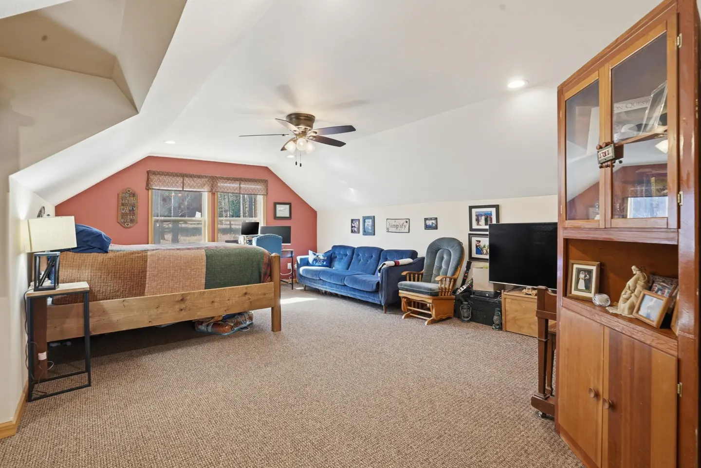 Spacious attic bedroom with a wooden bed, blue sofa, rocking chair, TV, and a tall wooden cabinet on carpeted flooring.
