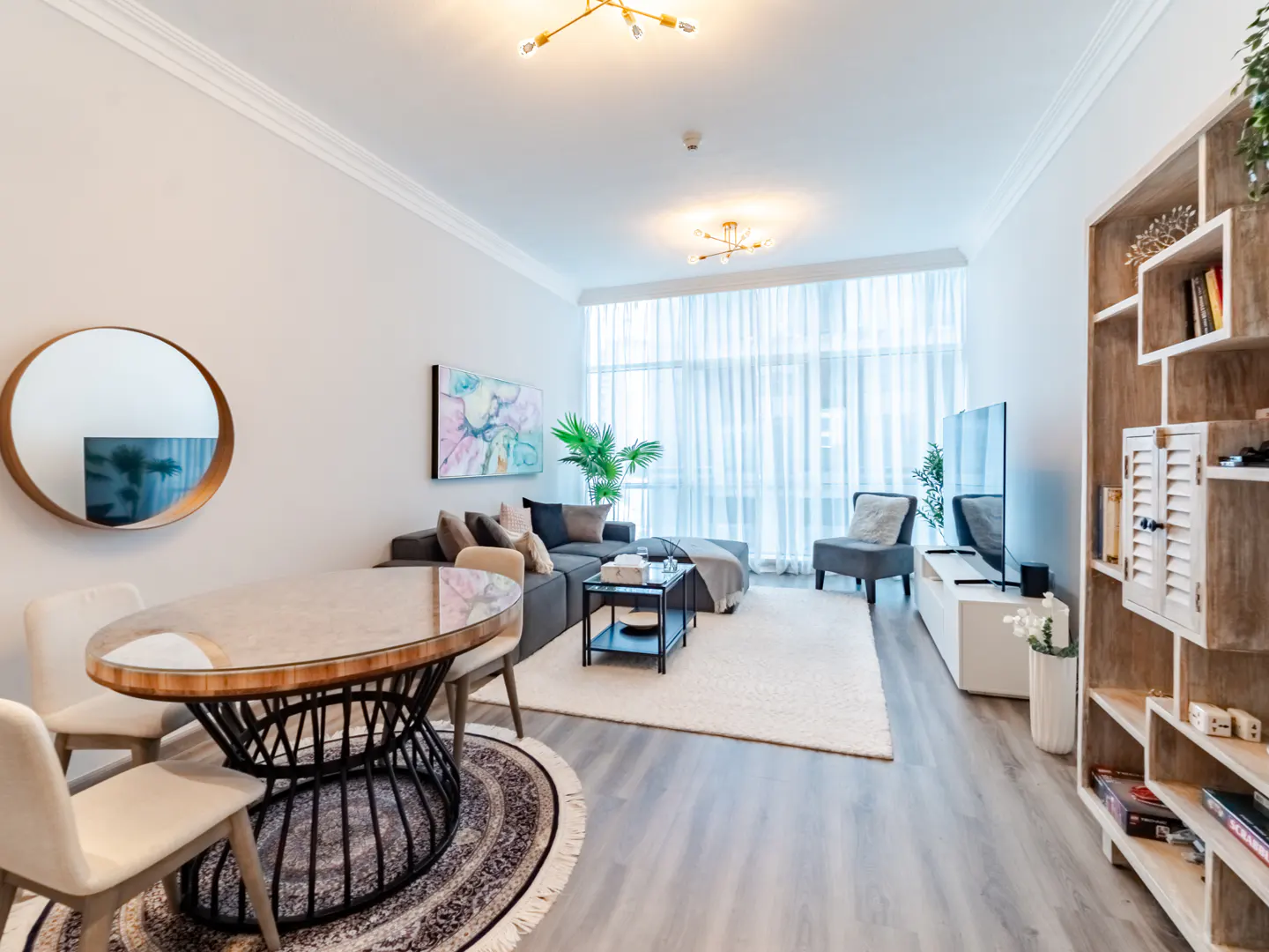 Bright, modern living room with gray sectional, oval table, and white rug. Natural light filters through sheer curtains. Bookshelf on the right.
