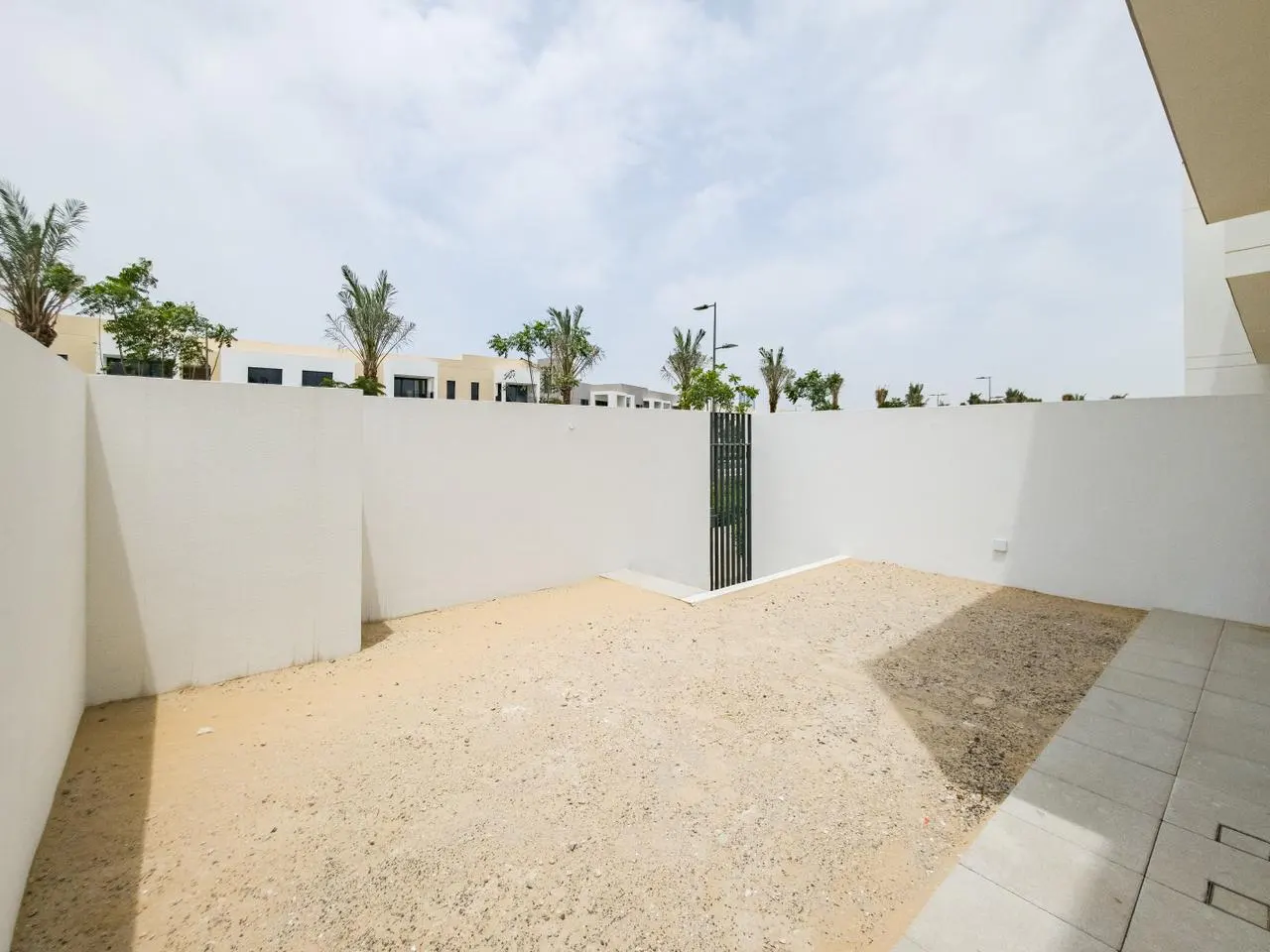 A backyard with a sand ground, surrounded by a white wall and a black metal gate. Palm trees and buildings are visible in the background.