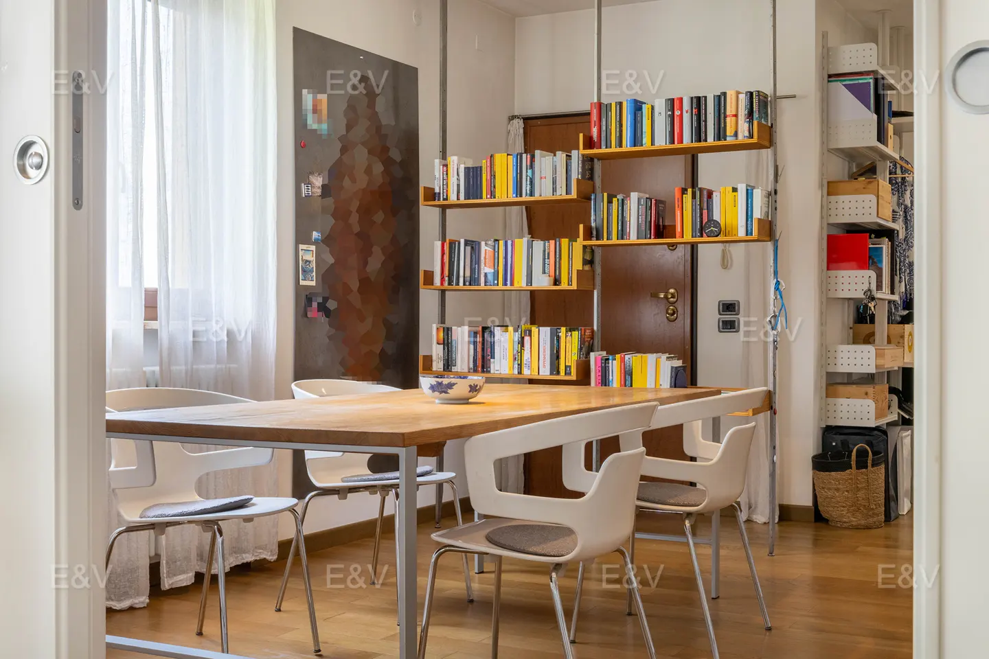 Dining area with a wooden table, four modern white chairs, and floor-to-ceiling shelving filled with colorful books.