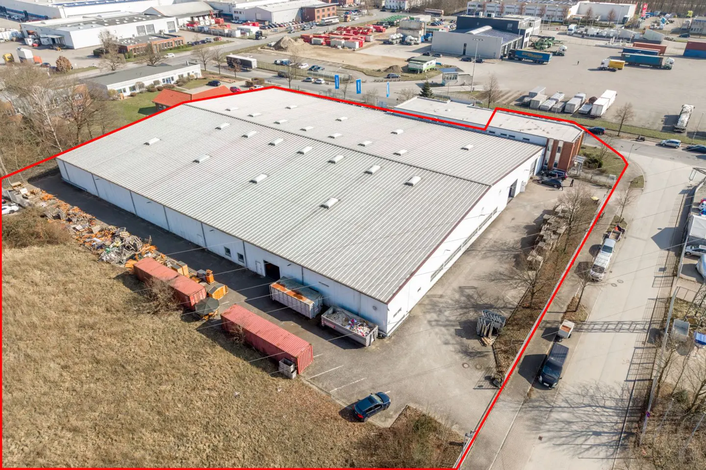 Aerial view of a large, white industrial building with a gray roof, outlined in red, surrounded by a paved lot and grassy areas.