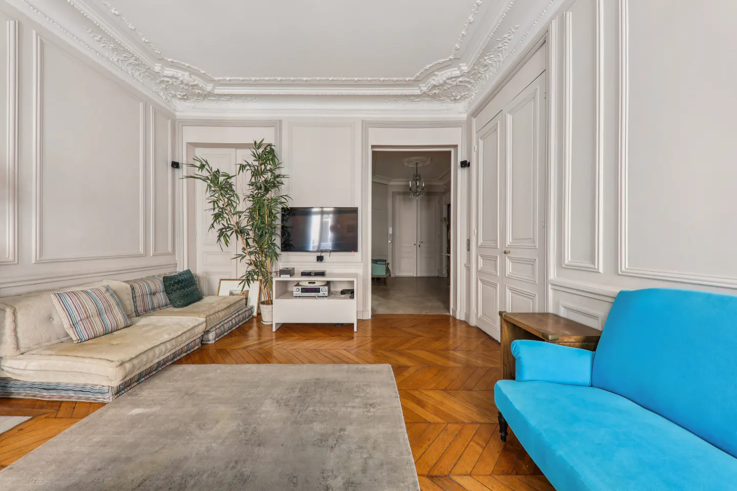 Bright living room with herringbone wood floors, white paneled walls, a beige floor sofa, and a vibrant blue armchair.
