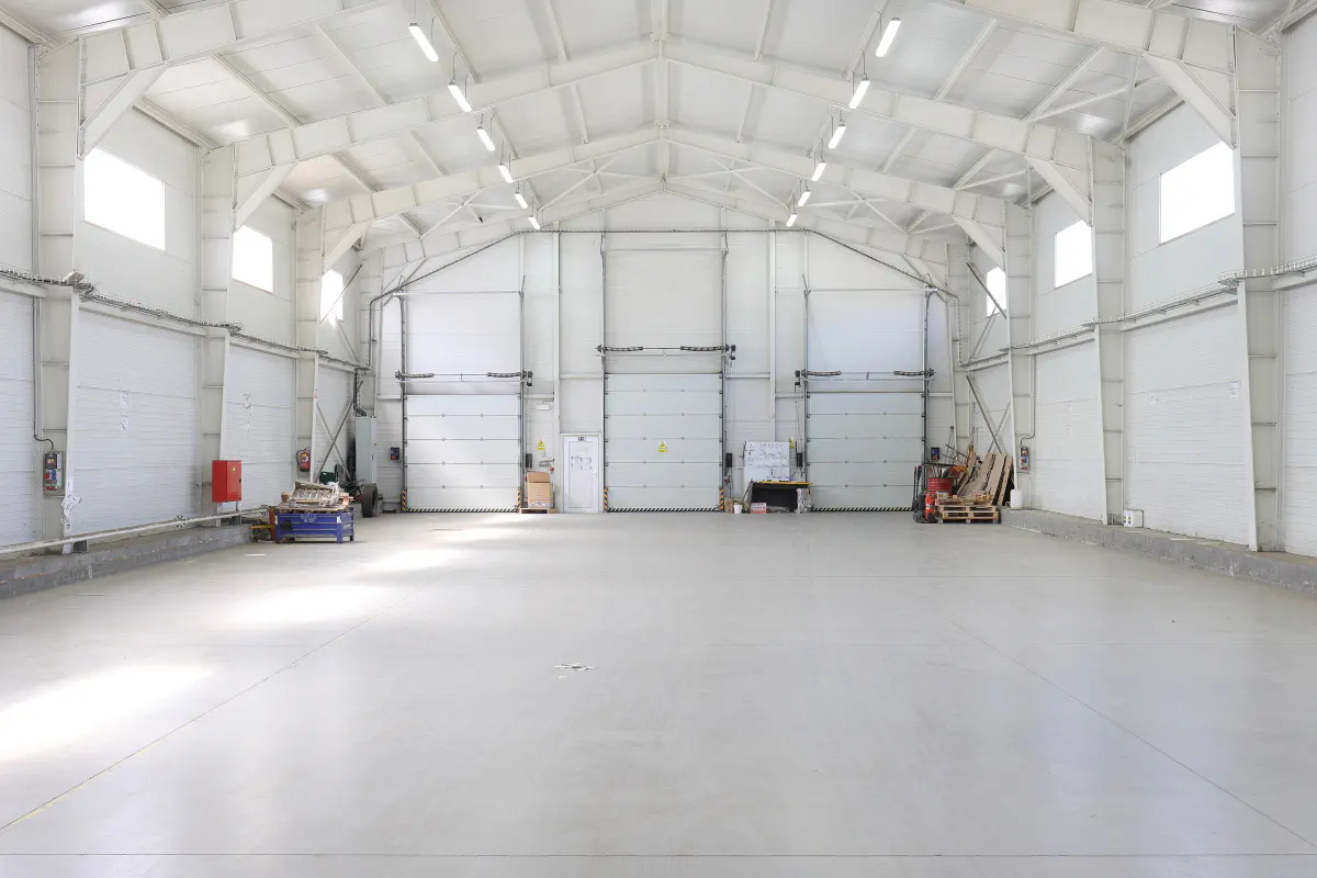 Bright, empty warehouse interior with white walls, concrete floor, and overhead lighting. Three closed garage doors are visible at the back.