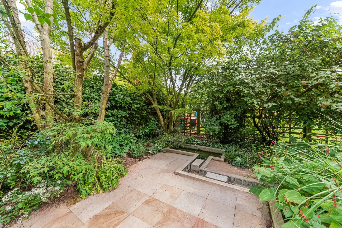A garden with a stone patio, green trees, bushes, and a small water feature. A fence is visible in the background.