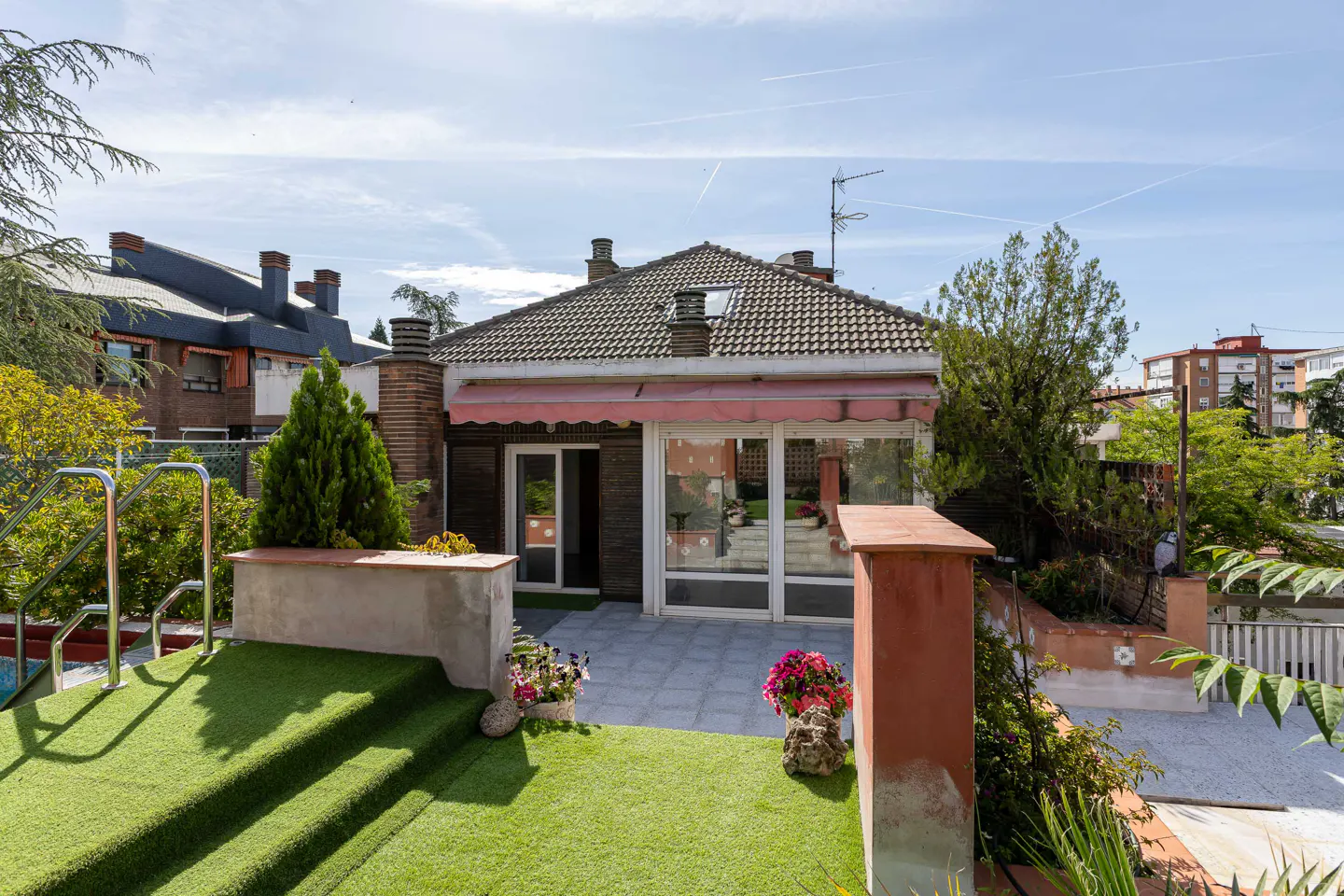 Backyard view of a single-story house with a gray tile roof, brick chimney, and a patio with glass doors and windows. Green grass and plants surround the patio.