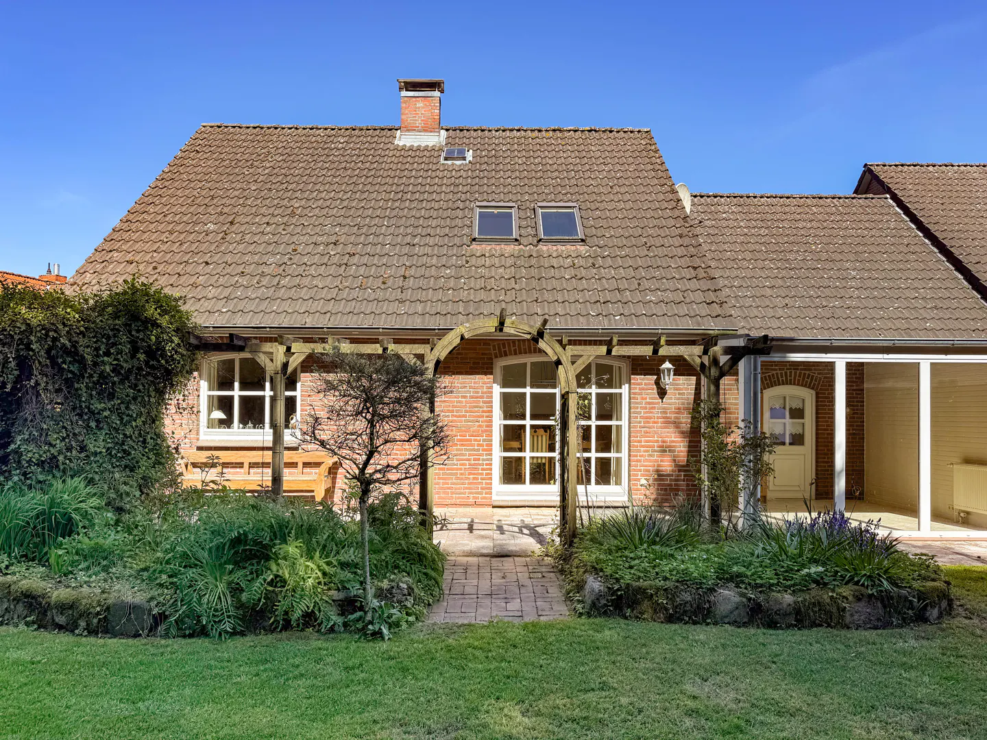 Backyard view of a brick house with a brown tile roof, a pergola, and a green lawn under a blue sky.