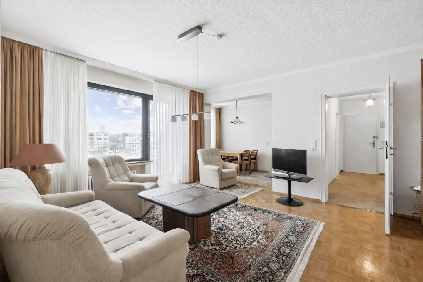 Bright living room with beige sofa, chair, and patterned rug. A dining area and TV are visible in the background. Natural light streams through a large window.