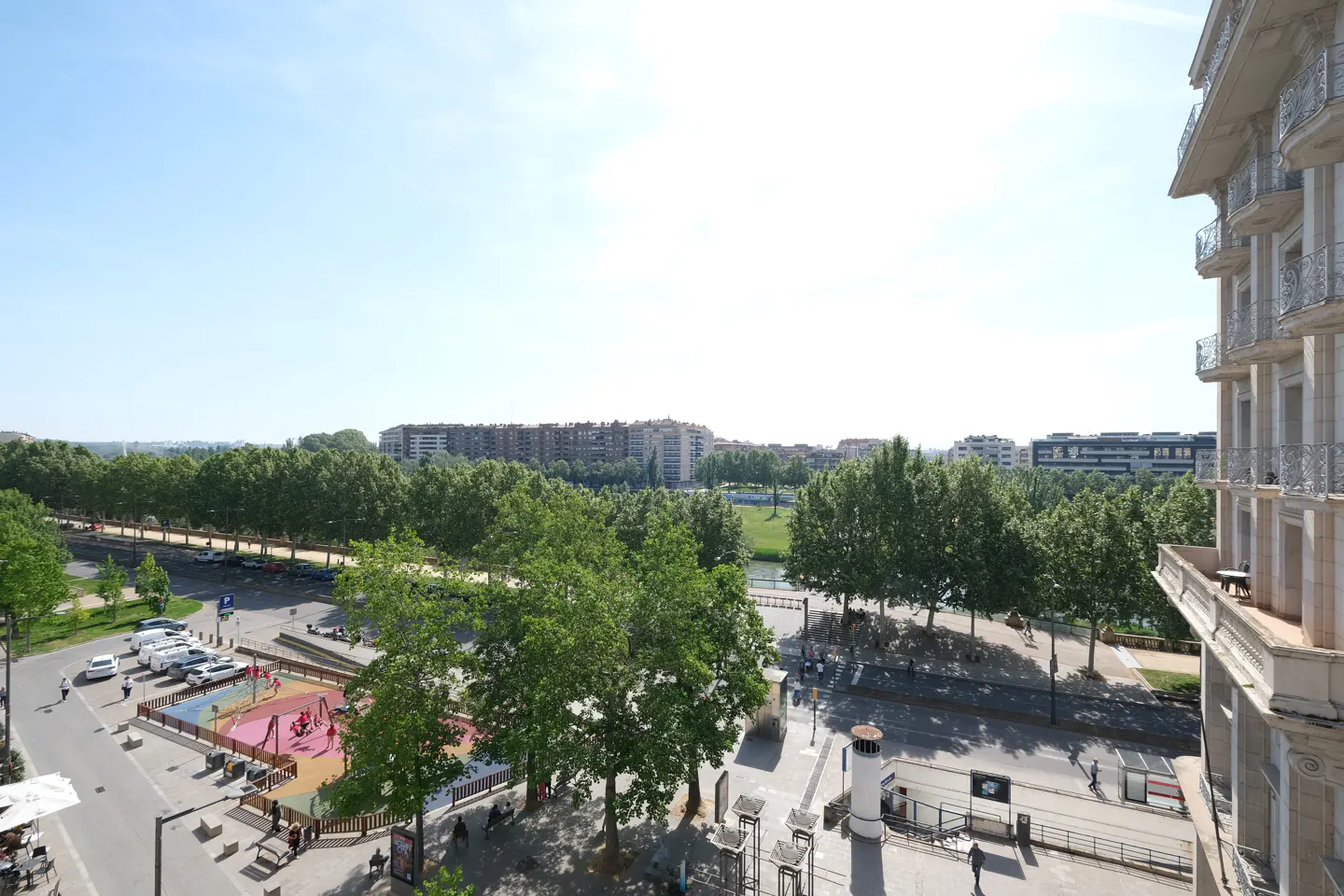 View from a balcony overlooking a city street with a playground, trees, and buildings under a bright blue sky.