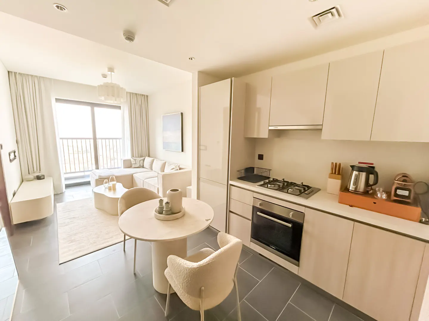 Modern open-plan apartment with a beige kitchen, round dining table, and a living area with a sofa and balcony access.