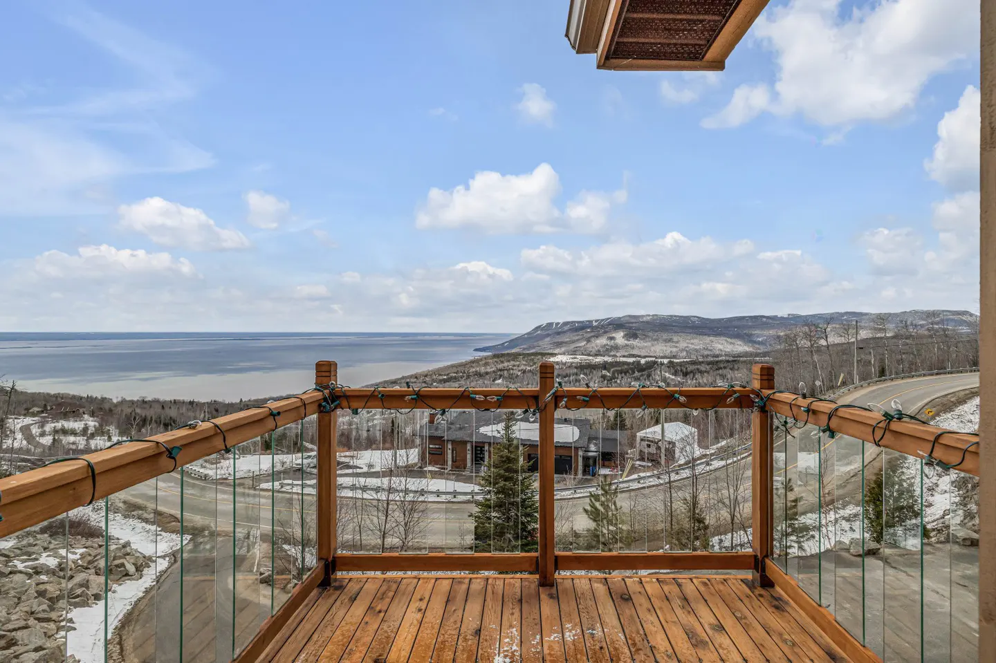 View from a wooden deck with glass railings, overlooking a bay, mountains, and a winding road on a partly cloudy day.