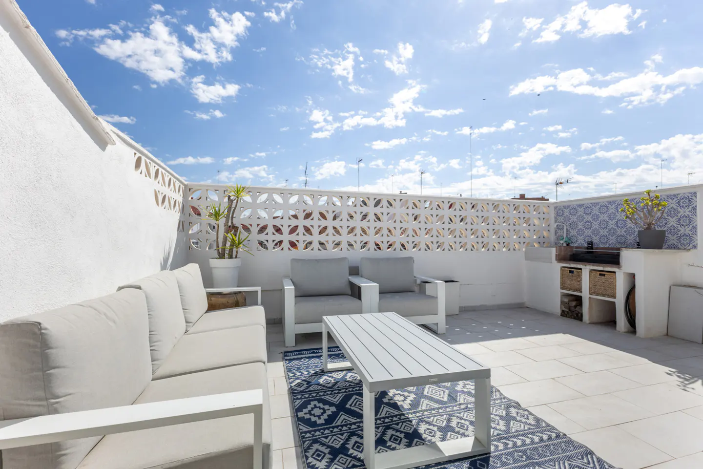 Bright rooftop patio with white furniture, blue patterned rug, and outdoor kitchen against a blue sky.