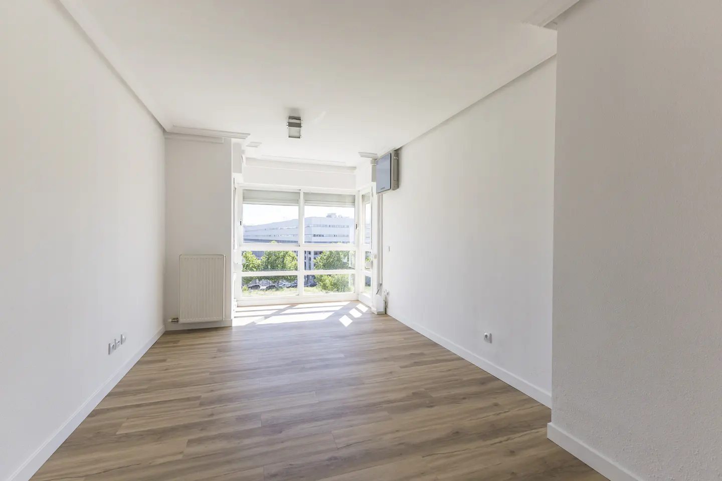 Bright, empty room with wood floors, white walls, and a large window with a view of trees and a building.
