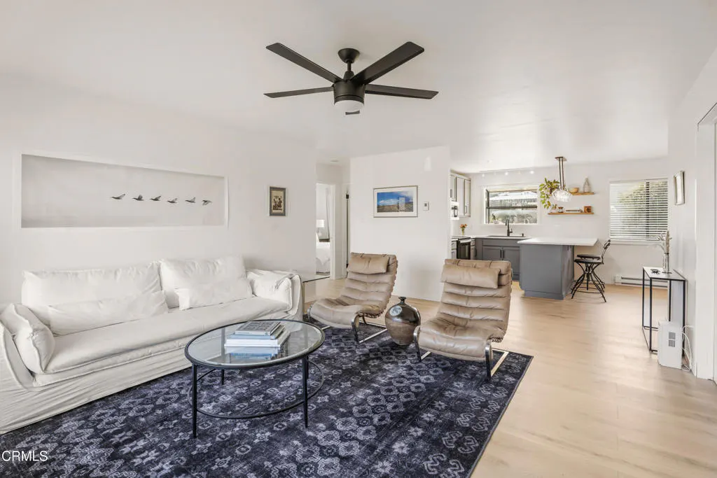 Bright living room with white walls, a white sofa, two leather chairs, and a blue patterned rug. A kitchen is visible in the background.