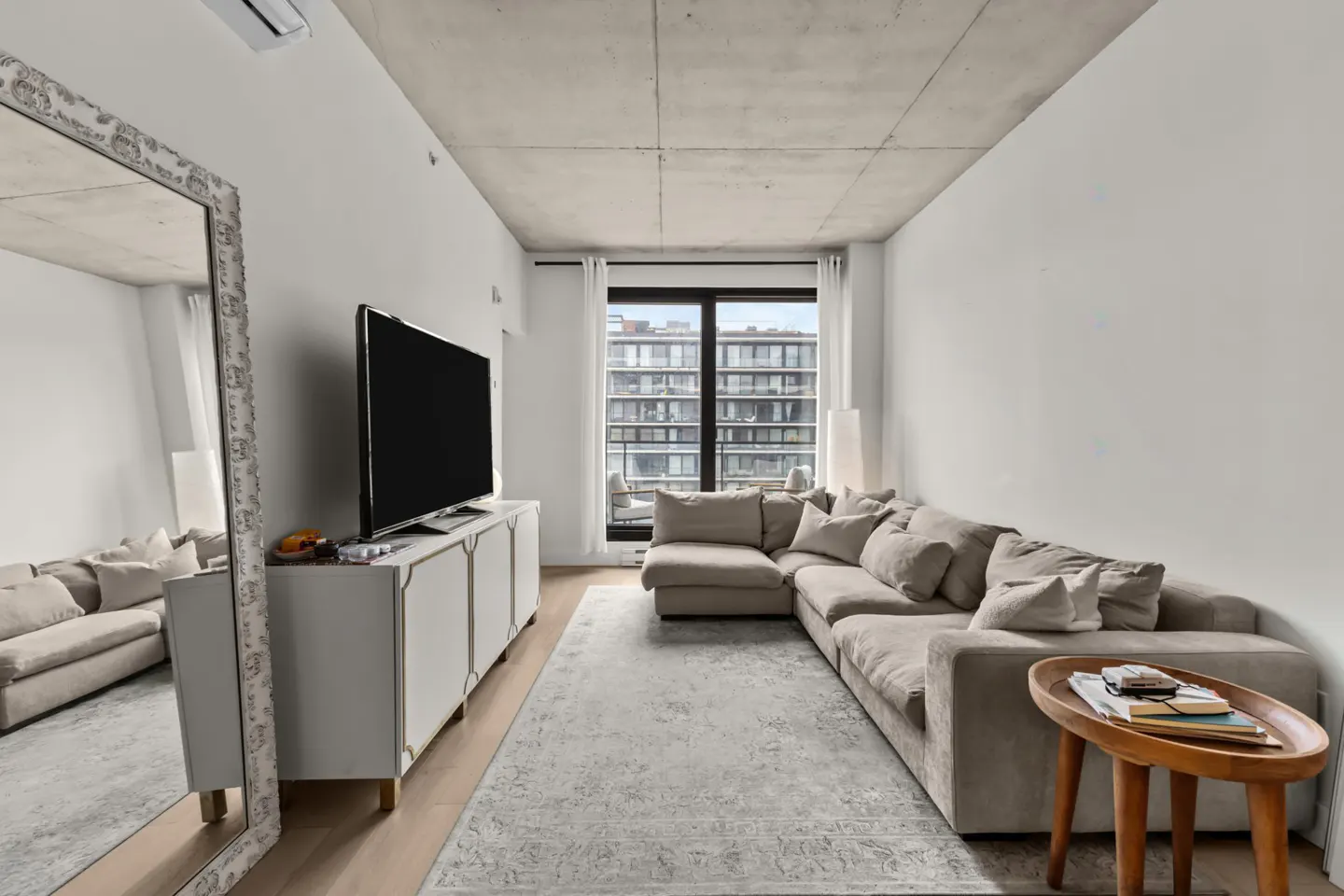 Living room with a beige sectional sofa, white media console with a TV, and a large mirror. A patterned rug covers the floor.