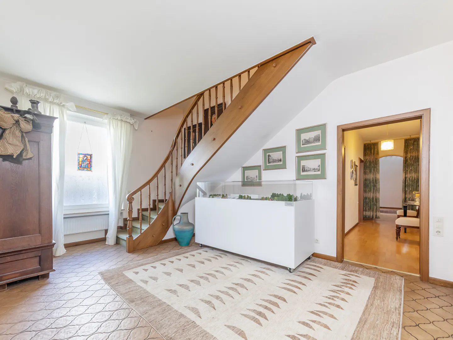 A bright room with a wooden staircase, a white display case, a patterned rug, and a doorway leading to another room.