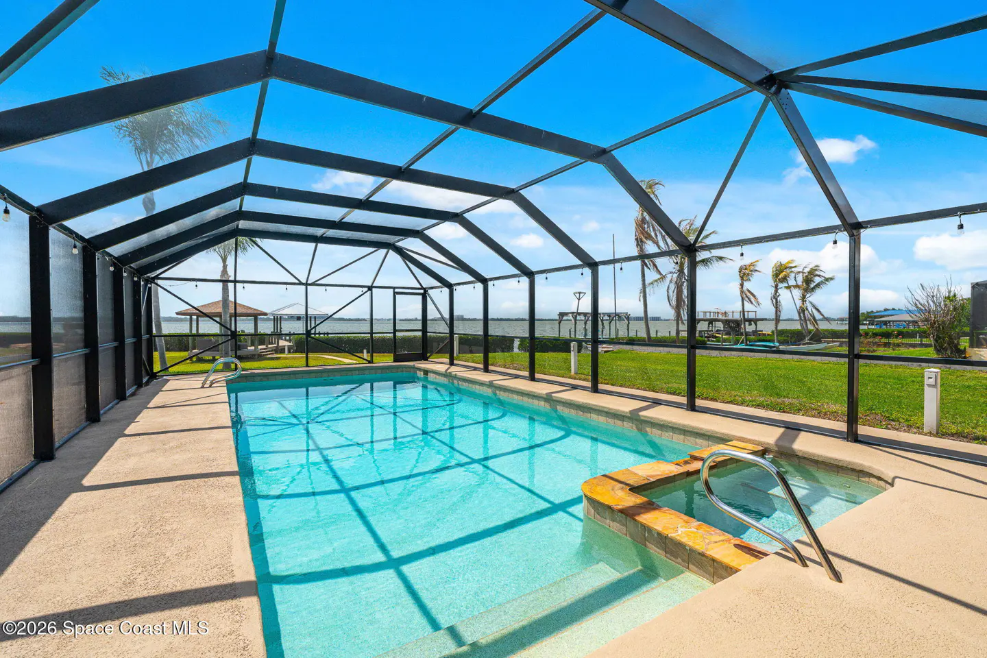 Screened-in pool with a hot tub. The pool is turquoise and the sky is blue. Palm trees and a gazebo are in the background.