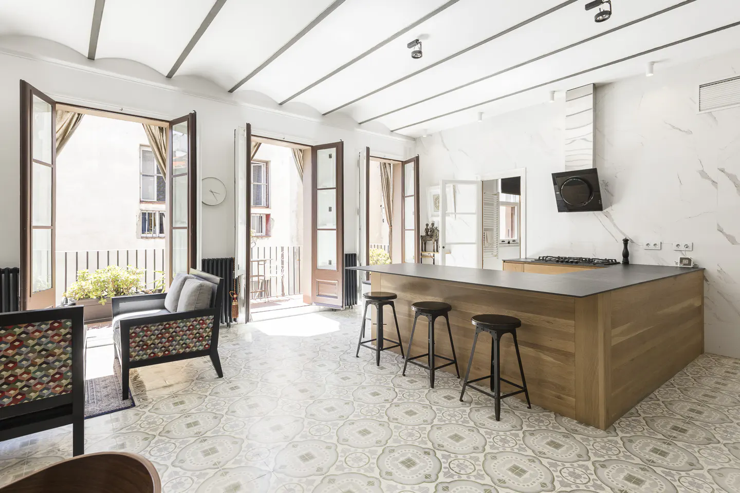 Bright, open kitchen with a wood island, black stools, and patterned tile floor. Balconies are visible through open doors.