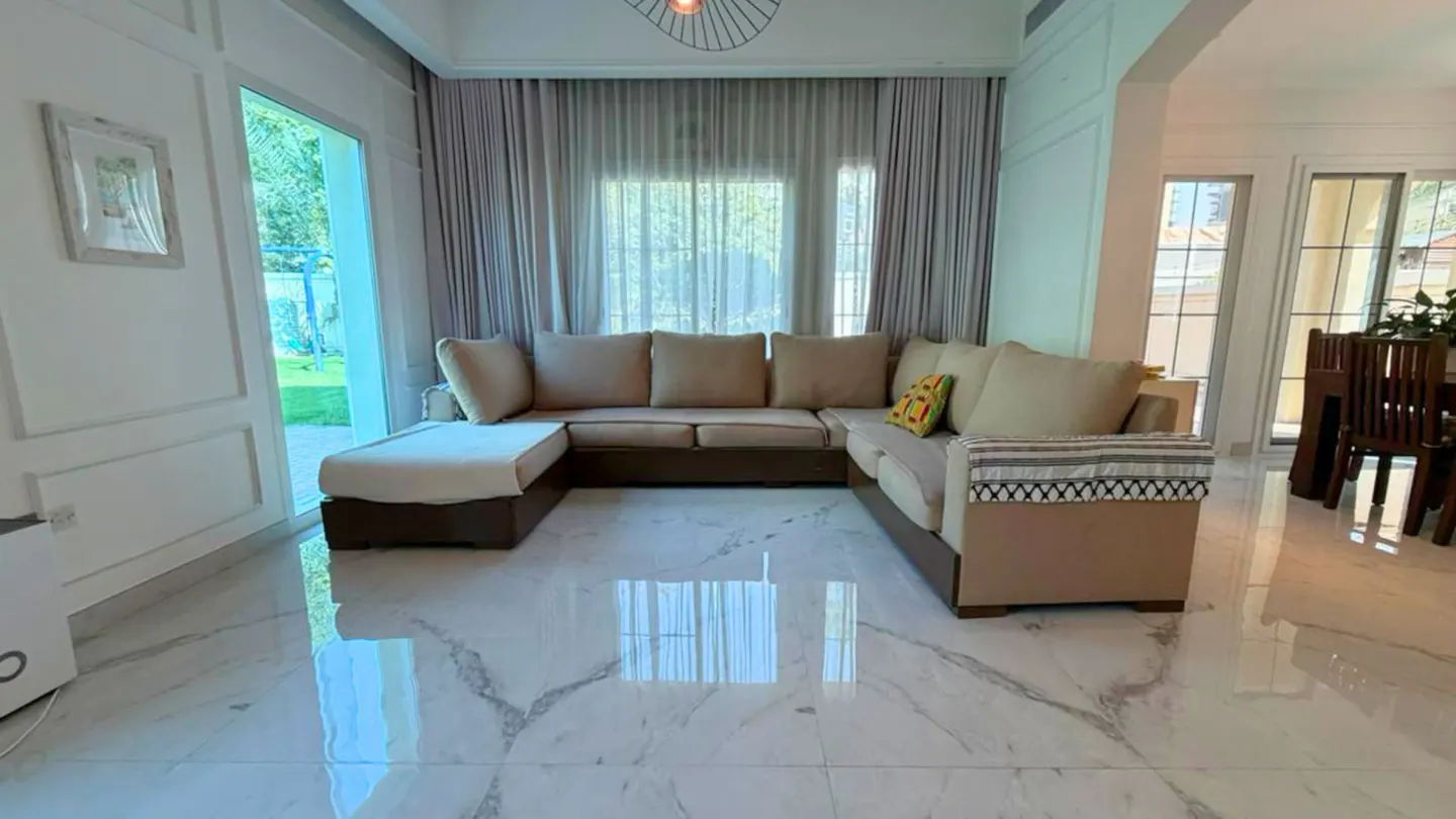 Living room with a beige sectional sofa, marble floors, and sheer curtains. A dining table is visible in the background.