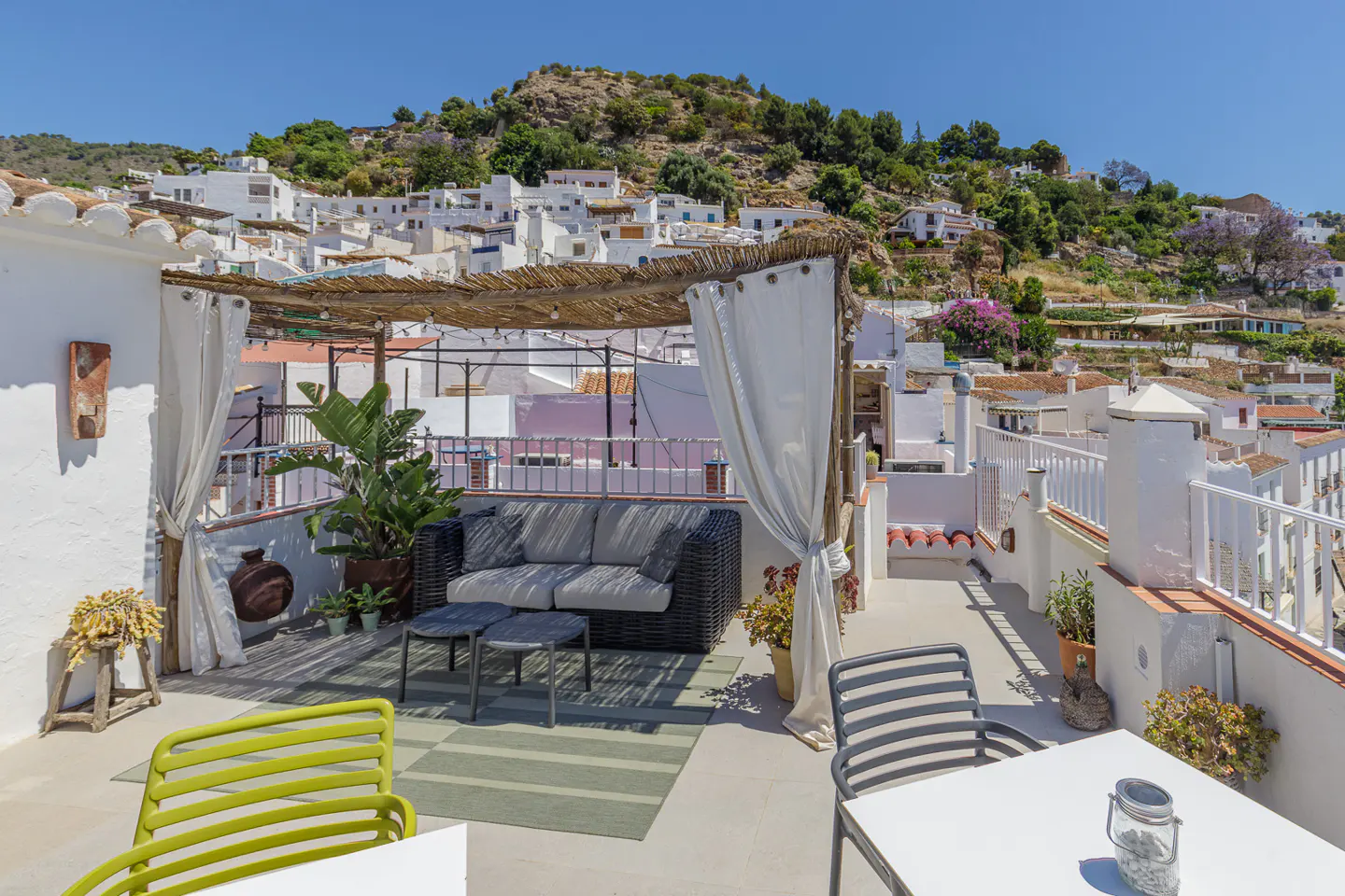 Rooftop terrace with a shaded lounge area, wicker sofa, and dining table, overlooking a hillside village of white houses.