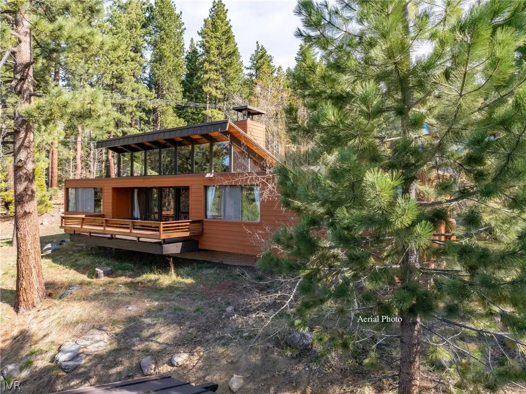 Exterior view of a modern, brown, two-story house with a deck, surrounded by tall green trees.