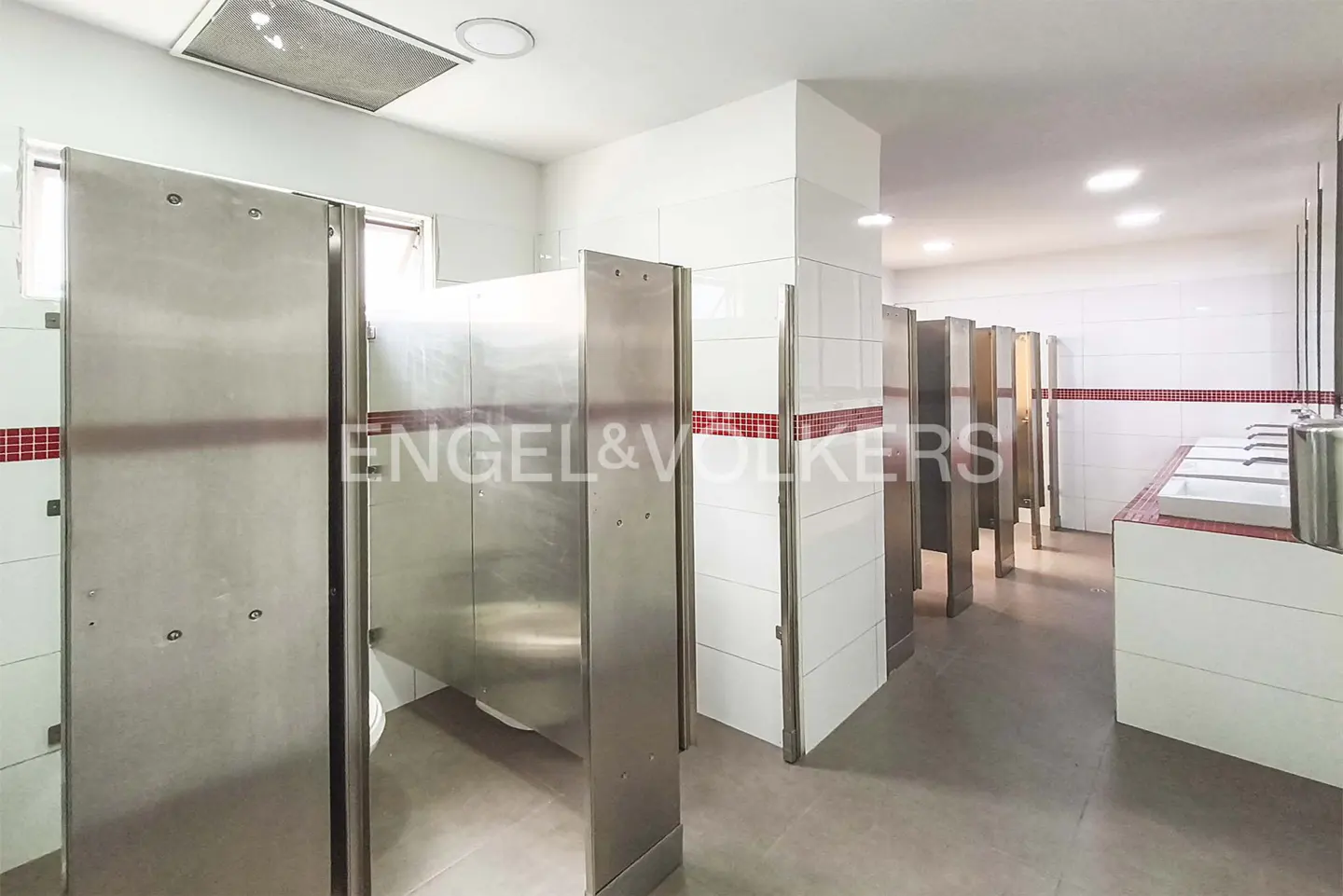 A commercial bathroom with stainless steel stalls, white tile walls, and red accent tiles.