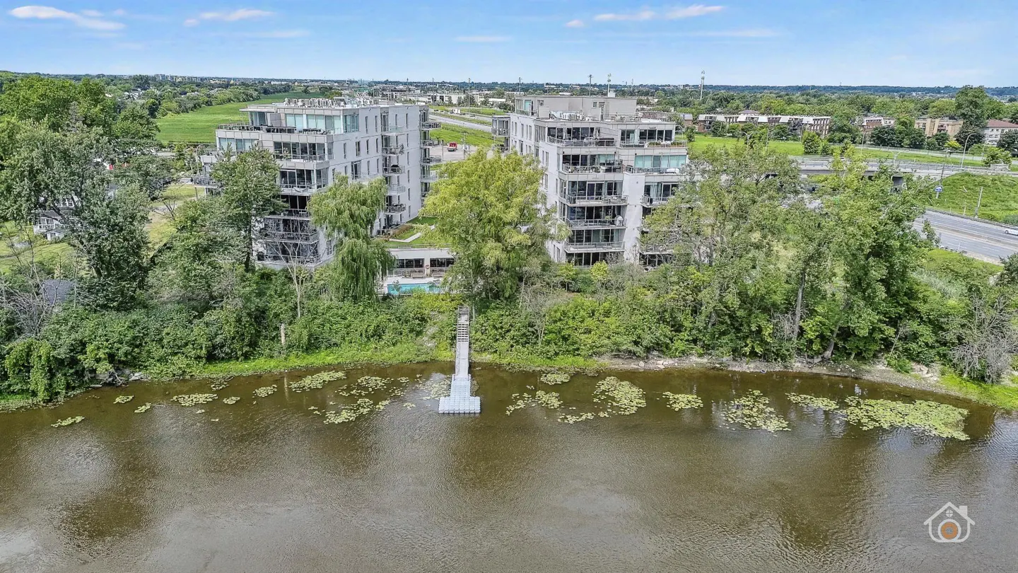 Aerial view of two modern, light gray condo buildings with balconies, overlooking a lake with lily pads and green trees.