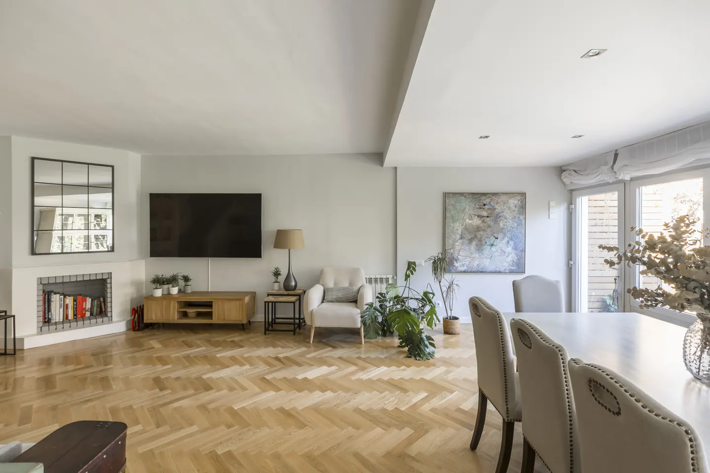 Bright, open-plan living and dining area with herringbone wood floors, fireplace, TV, and large windows.