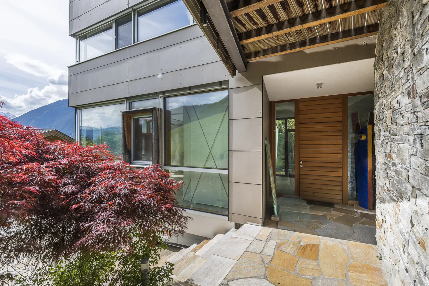 Modern home exterior with stone steps leading to a wooden door. A red Japanese maple tree is on the left. Large windows reflect the mountain view.