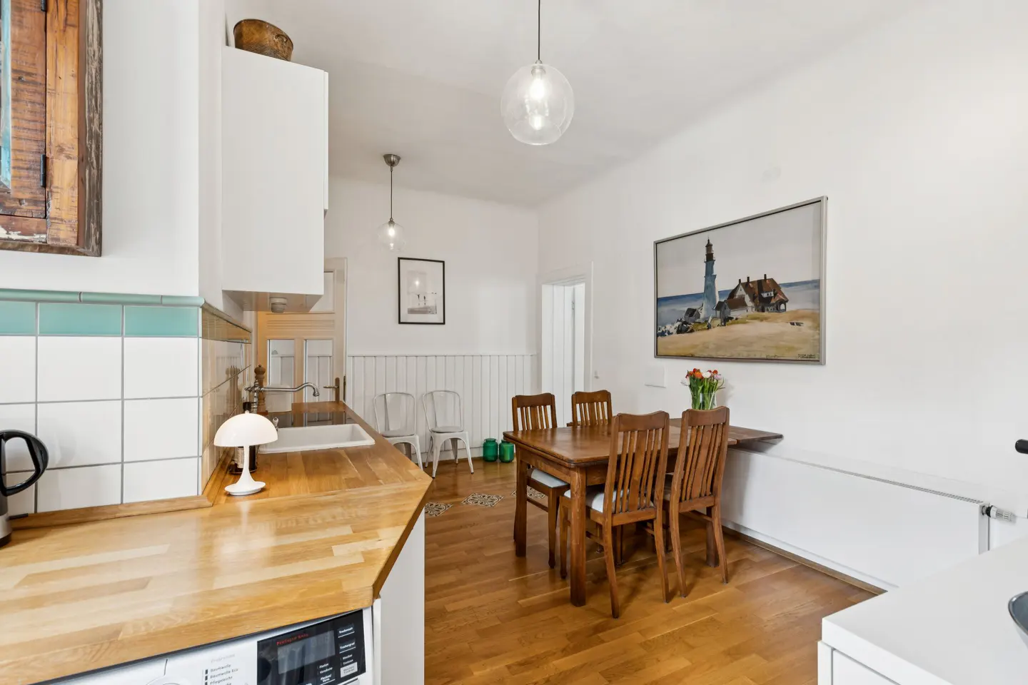 Bright kitchen with wood floors, white walls, and a wooden dining table with four chairs. A painting of a lighthouse hangs on the wall.