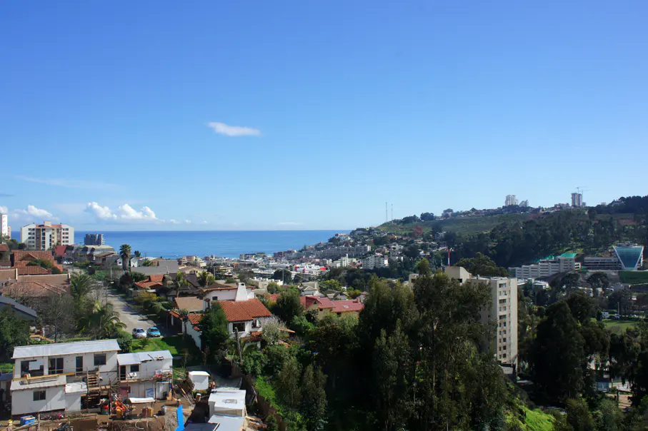 Cityscape view with red-roofed houses, green trees, and the ocean under a blue sky.