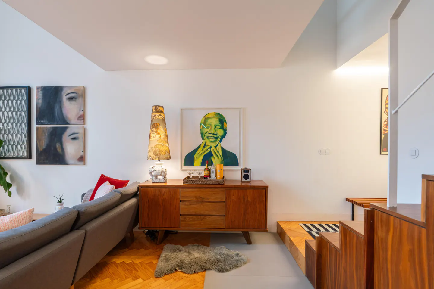 Living room with gray sofa, wood cabinet with Mandela art, patterned lamp, and stairs to another level.