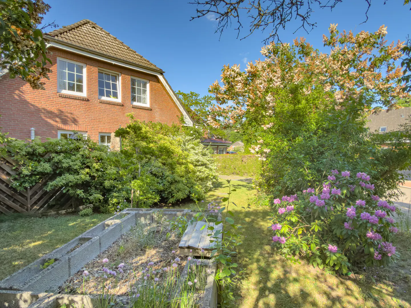 Exterior view of a red brick house with a garden featuring a raised bed, flowers, and lush greenery.