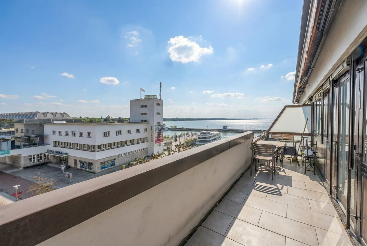 Balcony view of a white building, a pier, and a body of water under a blue sky. Table and chairs on the balcony.