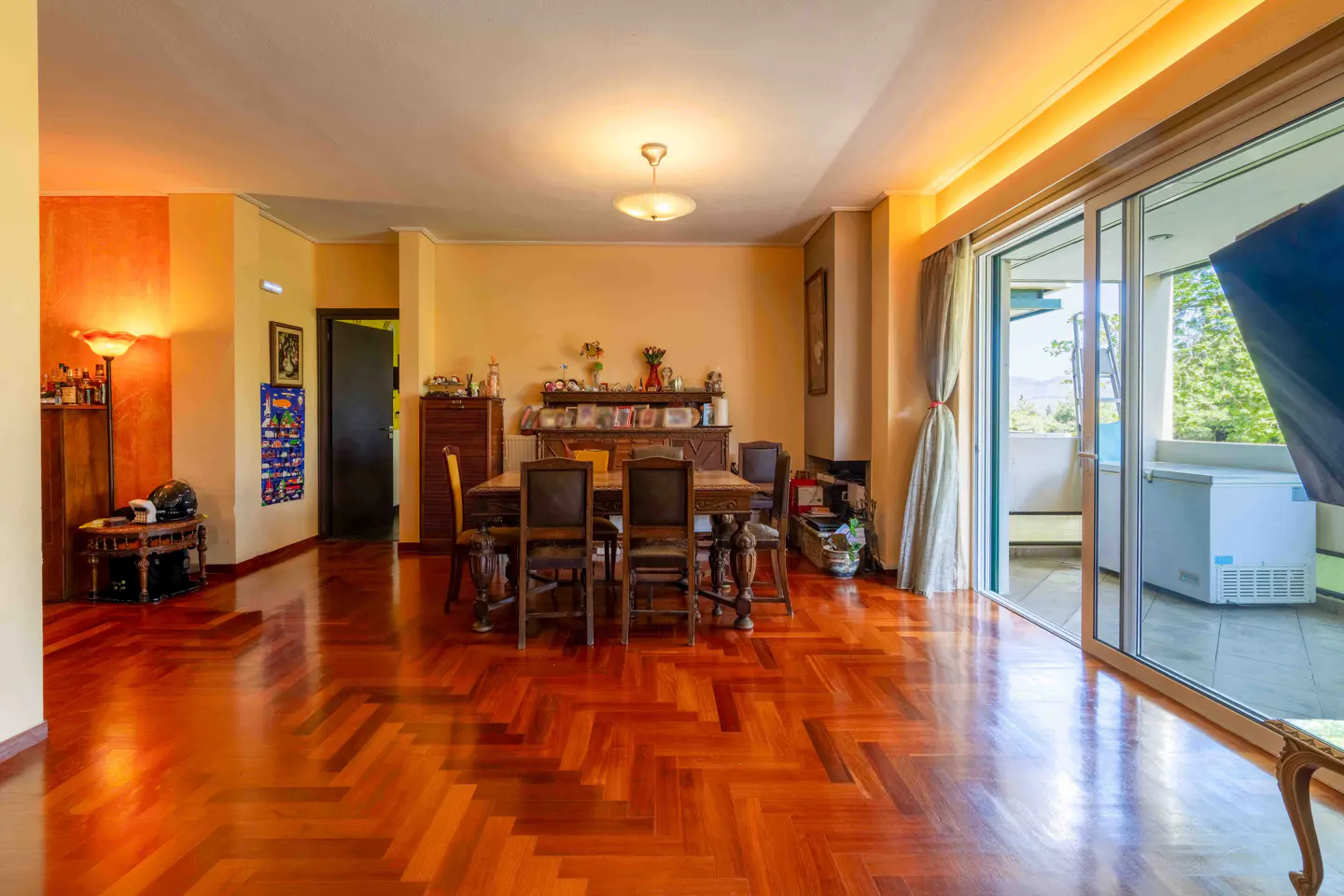 A dining room with a wooden table and chairs, a shiny herringbone floor, and a balcony with a view.