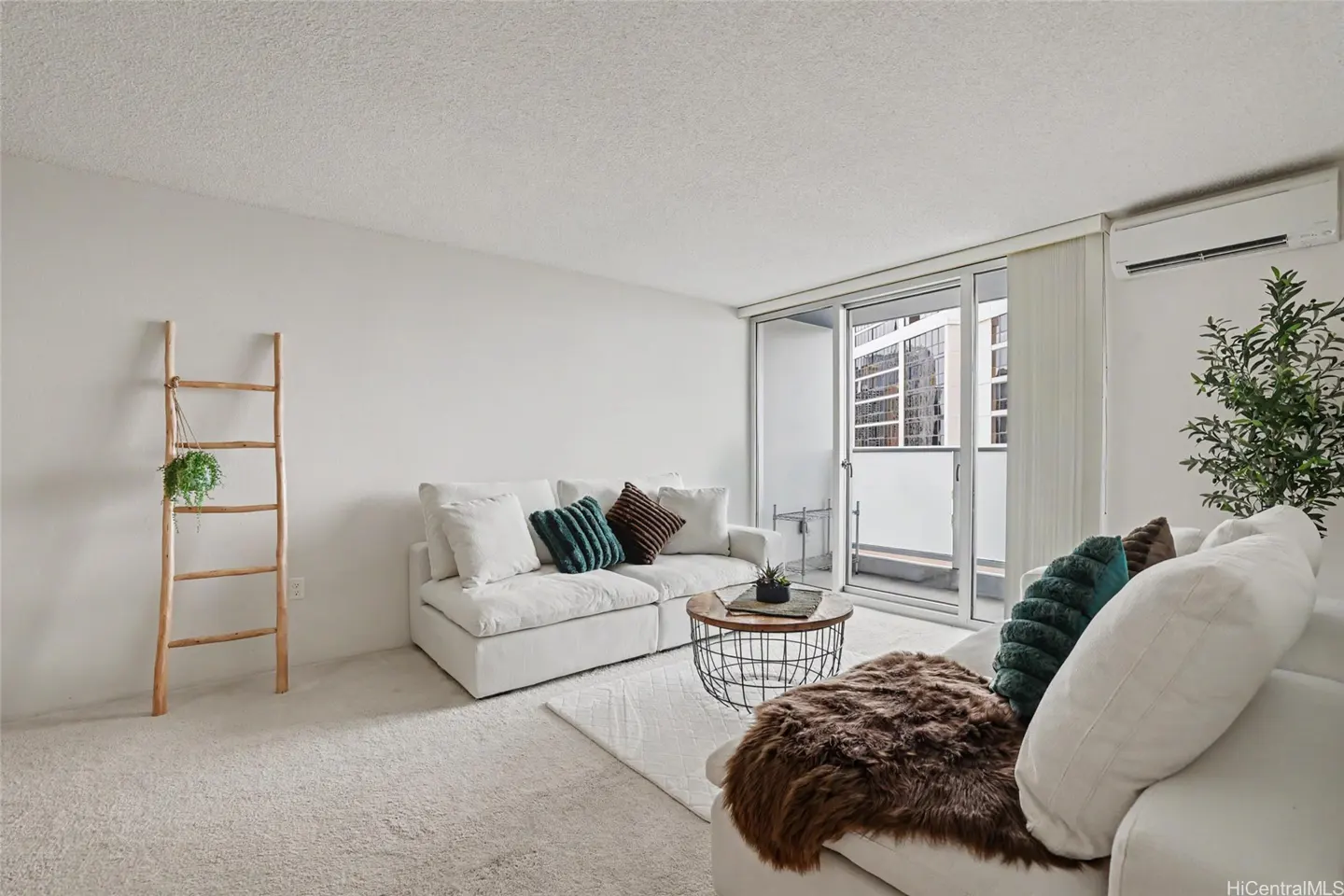 Bright living room with white walls, carpet, and sofas. A wooden ladder with a plant hangs on the left wall. Sliding glass doors lead to a balcony.