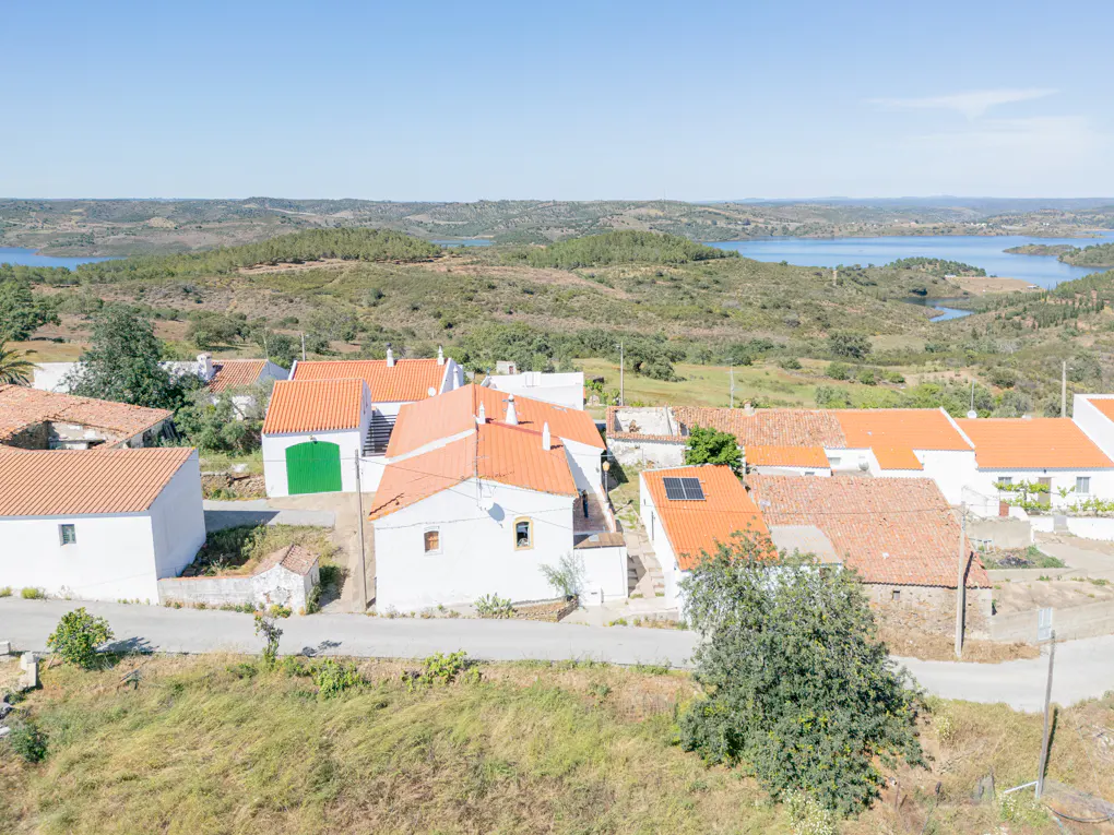 Aerial view of a rural village with white houses and orange roofs, set against a backdrop of rolling hills and a blue lake.