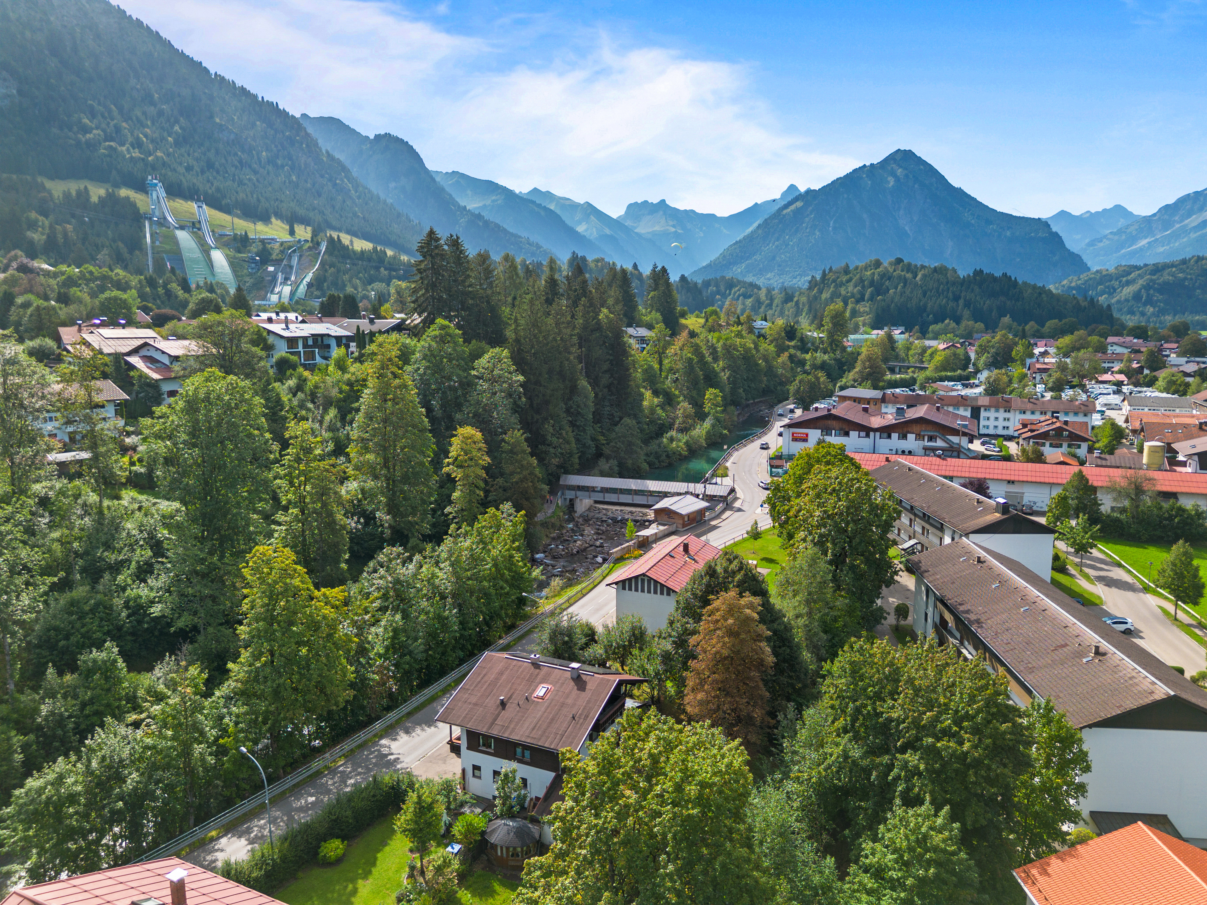 Aerial view of a town nestled in a lush green valley surrounded by towering mountains under a bright blue sky. [AI-generated text]