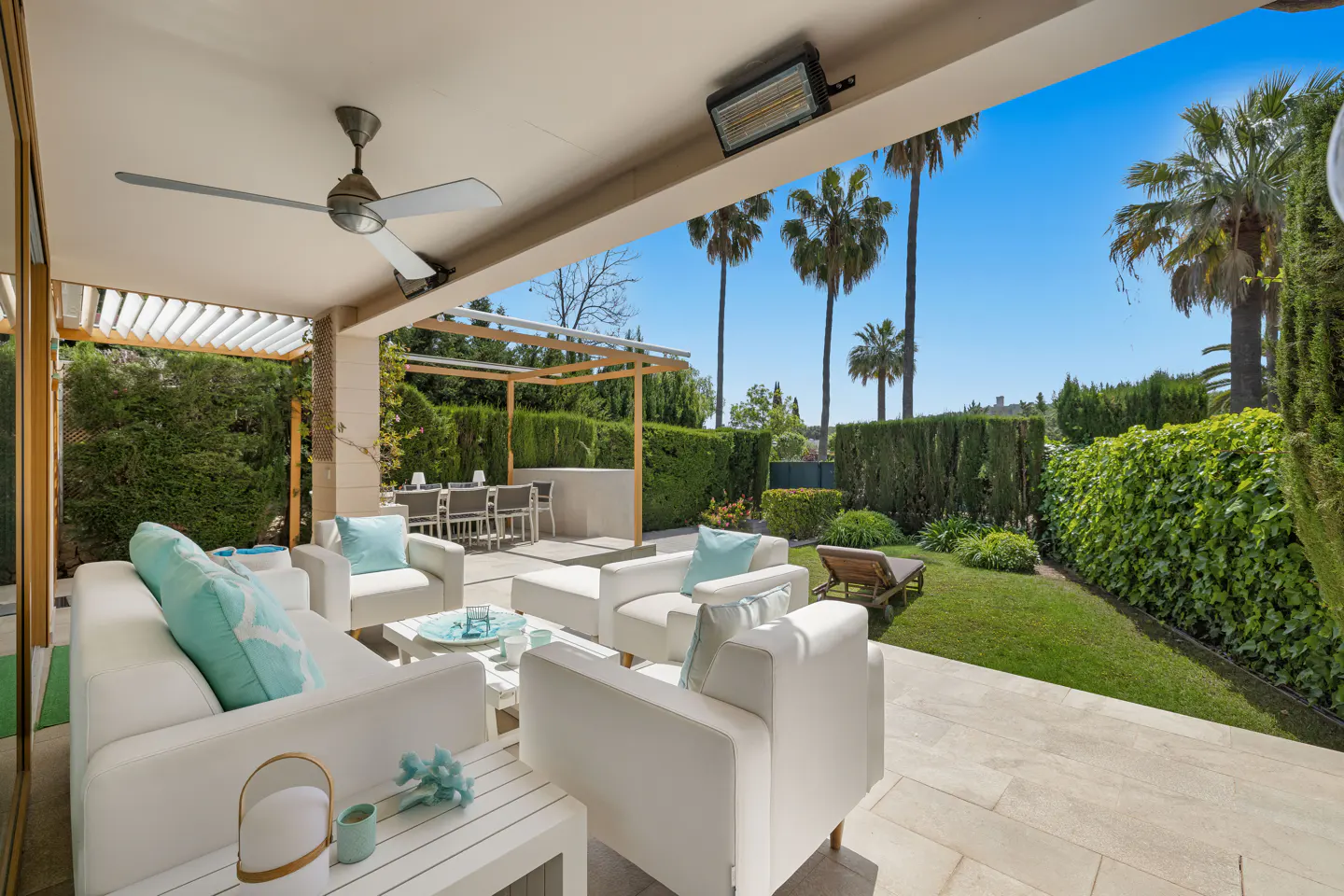 Outdoor patio with white furniture, turquoise pillows, and a ceiling fan. Palm trees and green hedges in the background.