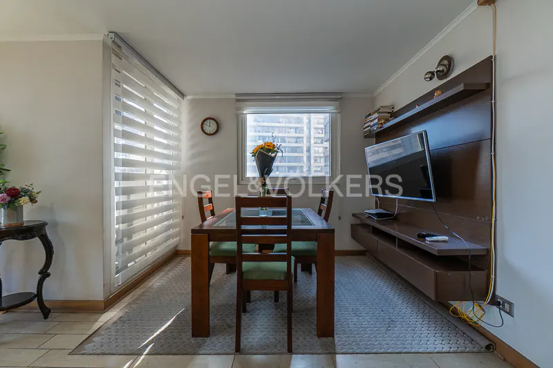A dining room with a wooden table, green chairs, and a TV on a brown wall unit. A window with blinds is in the background.