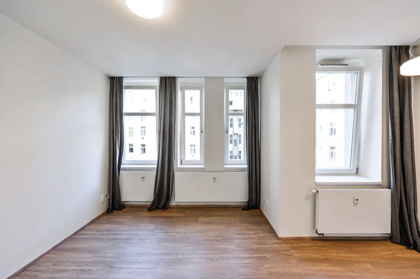 Bright, empty room with wood floors, white walls, and three windows with gray curtains. A white radiator is under one window.