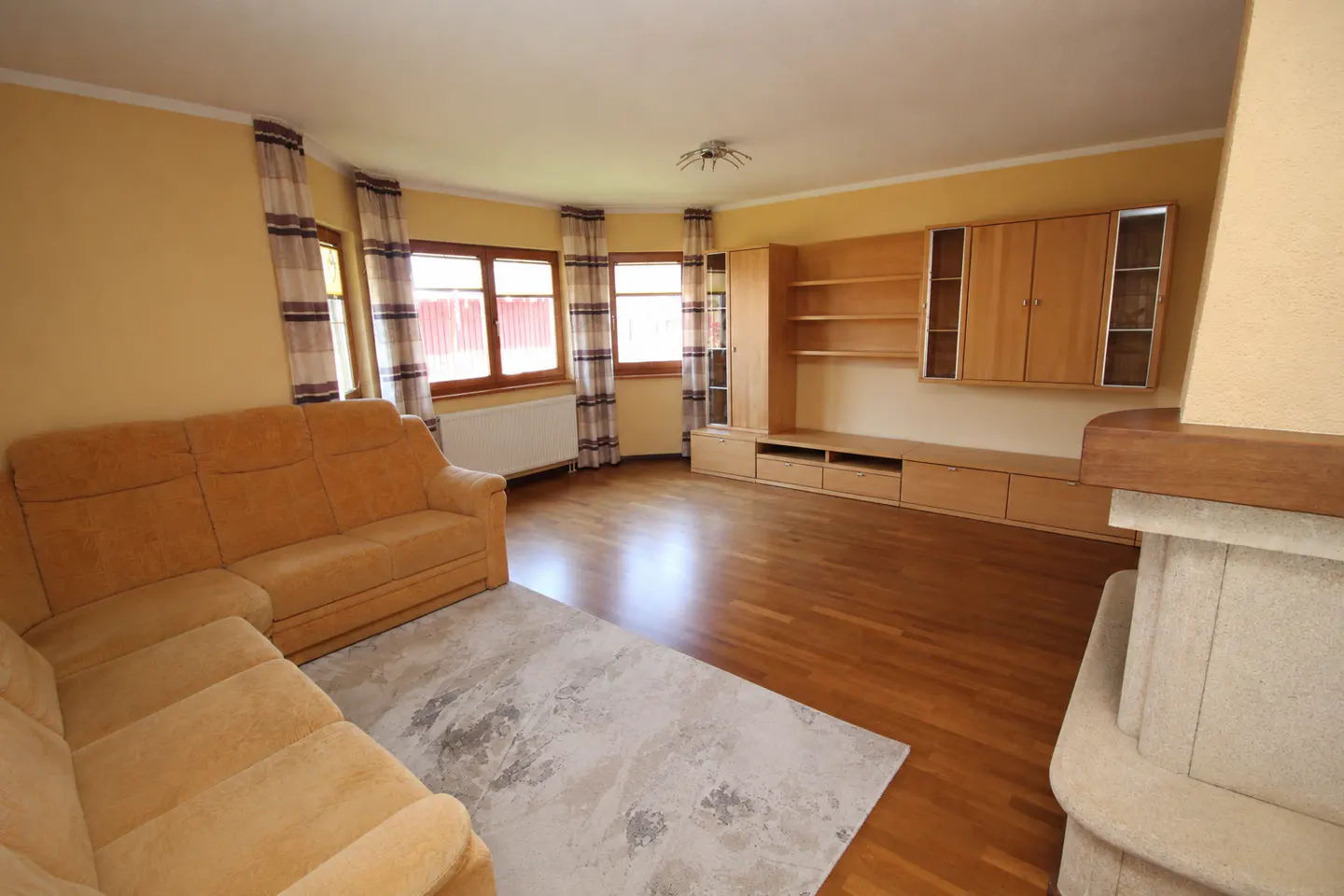 Living room with yellow walls, wood floors, and a tan sectional sofa on a gray rug. Windows with striped curtains and a built-in wooden entertainment center.