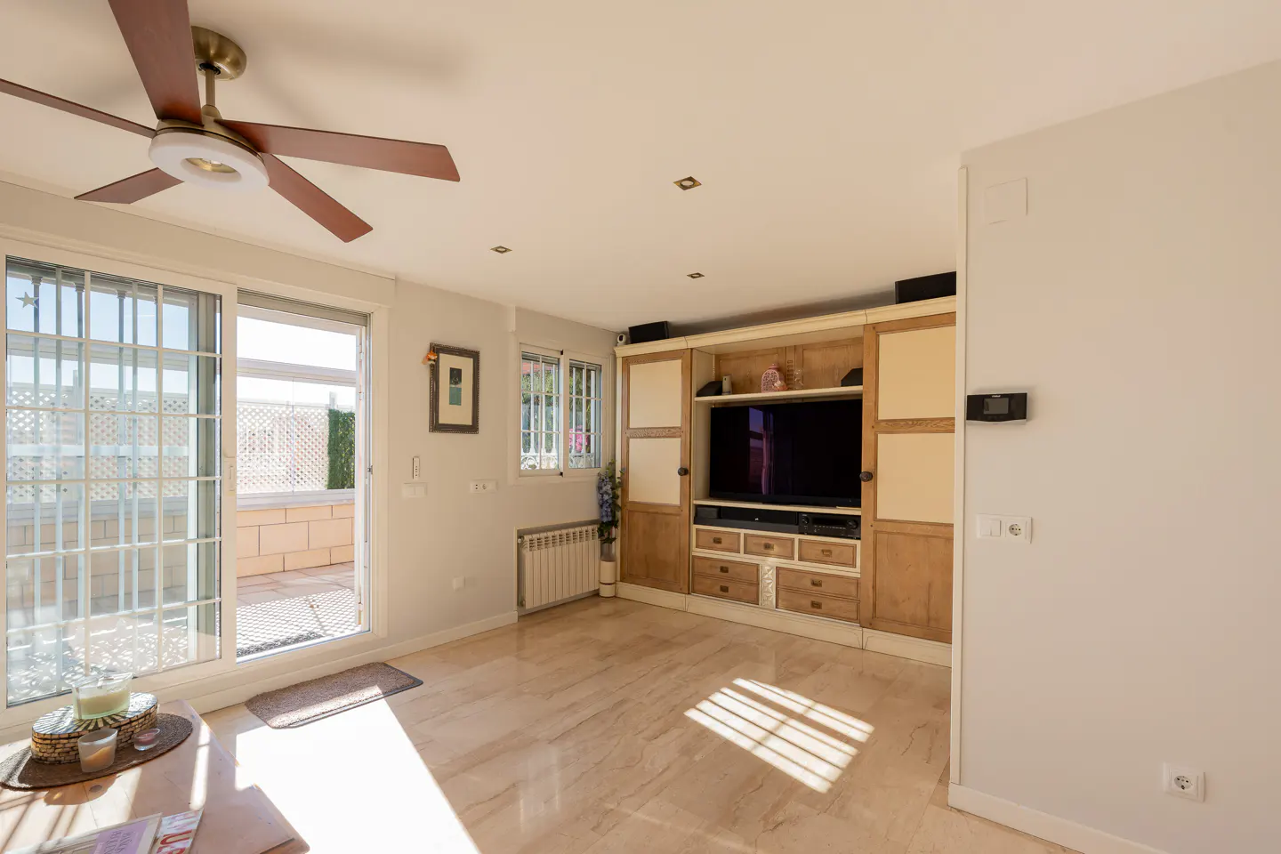 Bright living room with marble floors, a ceiling fan, and a built-in entertainment center with a large TV. Sliding glass doors lead to a sunny patio.