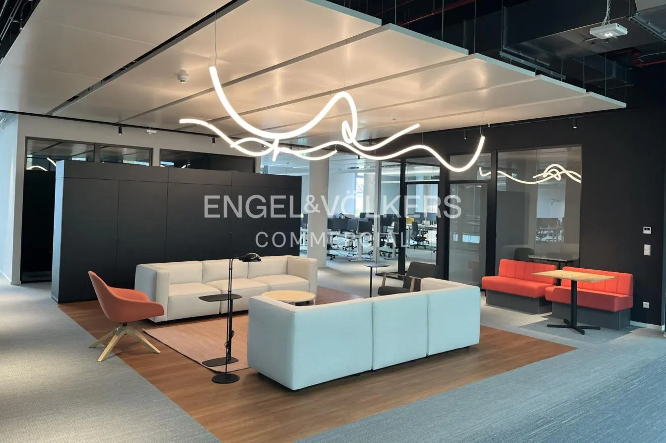Modern office space with white sofas, orange chair, and unique lighting fixture. Engel & Volkers logo visible.
