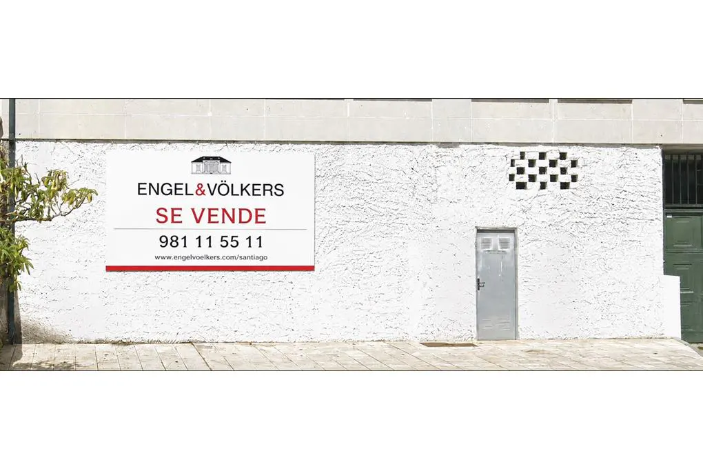 Engel & Völkers real estate for sale sign on a white textured wall with a small metal door and a tree on the left.