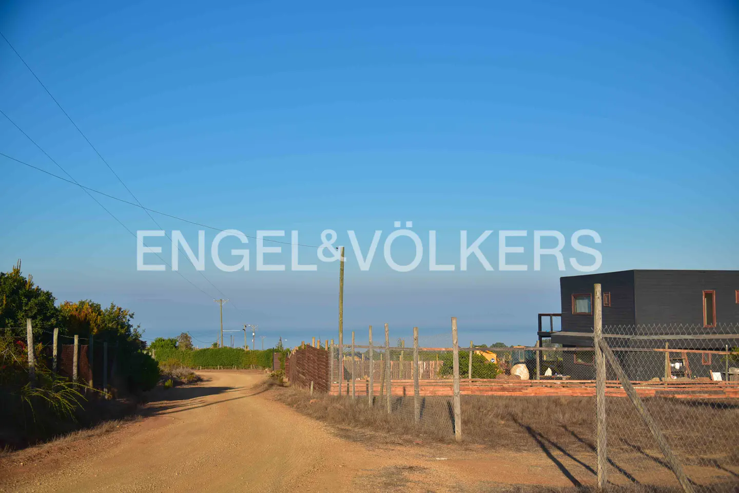 Dirt road leads to a modern black house with a fence, under a clear blue sky. Engel & Völkers logo is superimposed.