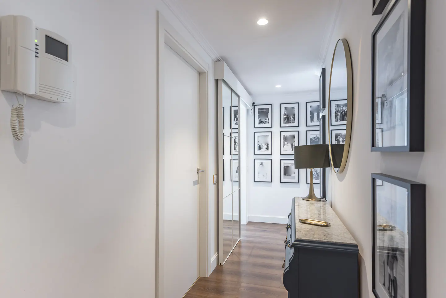 Hallway with white walls, wood floors, and black and white framed photos. A dark blue dresser with a lamp and gold mirror are on the right.