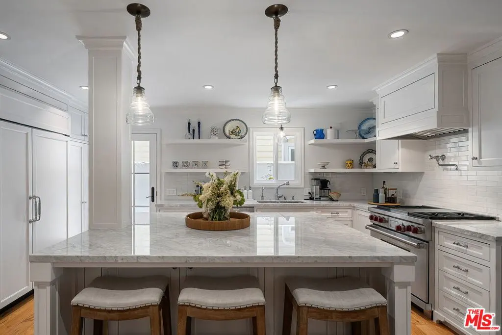 Bright, white kitchen with marble island, three stools, and pendant lights. Open shelves display decorative items. Stainless steel appliances.