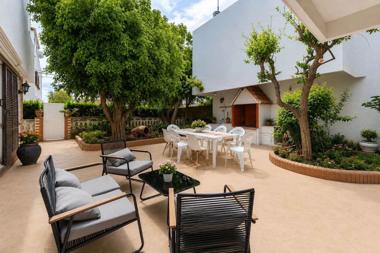 Outdoor patio with seating area, dining table, and grill. Lush green trees and plants surround the space. Neutral color scheme.