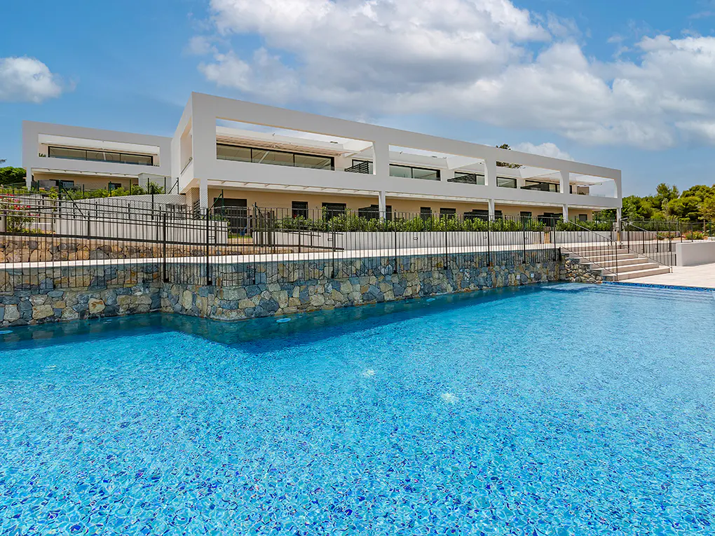 Modern white building overlooks a blue tiled pool. A stone wall and black fence separate the building from the pool. Blue sky.