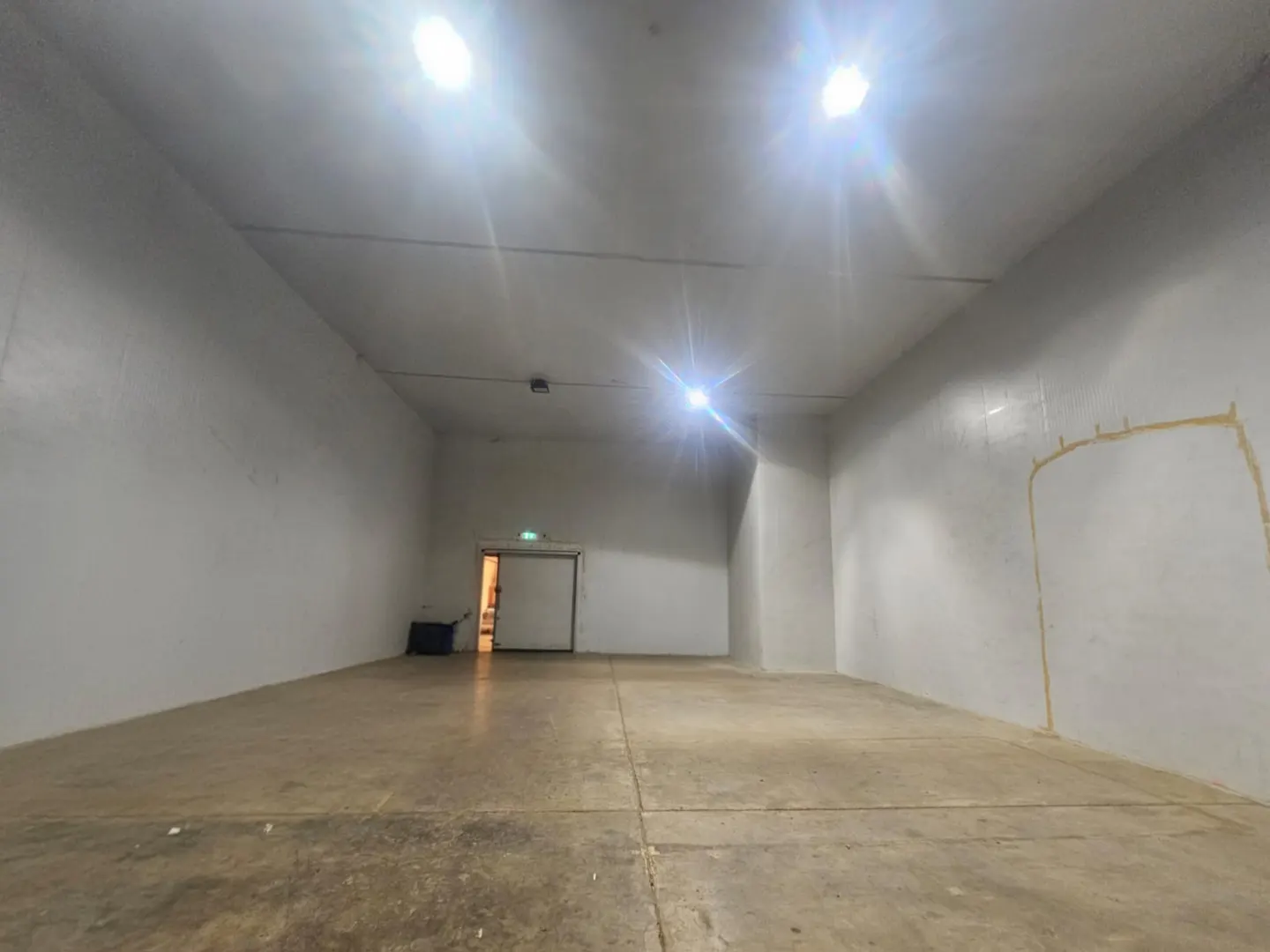 Empty warehouse interior with concrete floor, white walls, and bright overhead lighting. A doorway is visible in the background.